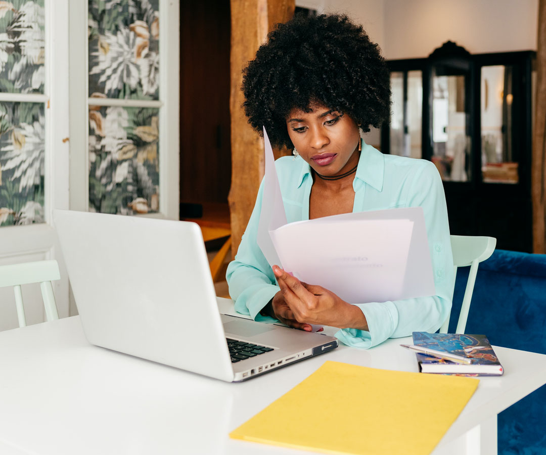 Person working on a laptop, surrounded by paperwork.